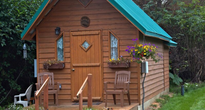 Wooden tiny home with a green roof. The home has flowers near the window and two chairs near the front door.