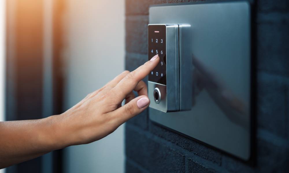 A hand reaches to type in a code on a home security system keypad on the exterior of a dark-brick home.