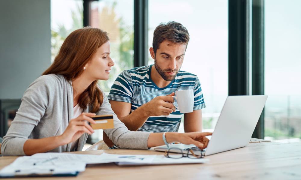 Two people look over a pile of papers and a computer, pointing at the screen while holding a credit card and coffee cup.