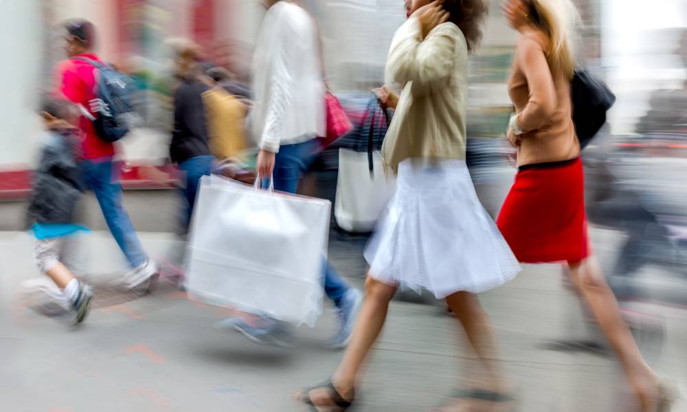 Several people walk by in a street in a fast fashion. They have shopping bags and seem to be in a city environment.