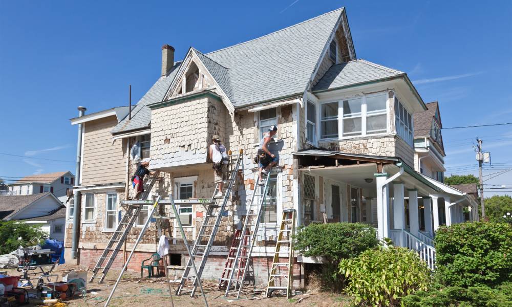 A lovely beach house undergoing renovations with multiple workers on ladders refurbishing the exterior.