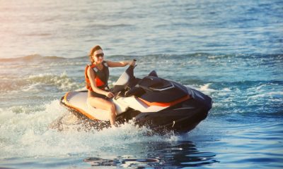 A woman wearing an orange life vest rides on the back of a large jet ski, creating waves on the water.