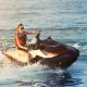 A woman wearing an orange life vest rides on the back of a large jet ski, creating waves on the water.