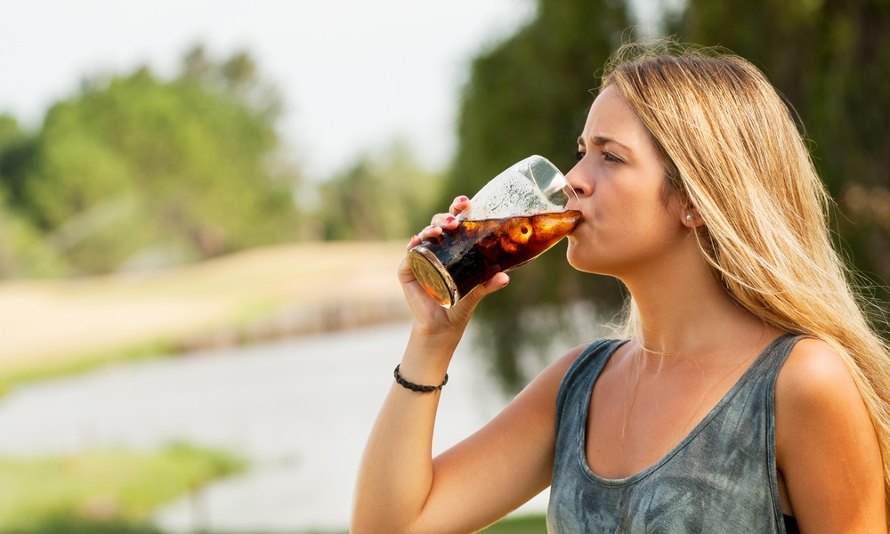 A blonde-haired woman wearing a tie-dye tank top drinking a glass of soda with ice while standing outside.
