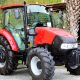 A red compact tractor sits on a gray concrete path. A large tree and a grassy area appear behind the tractor.