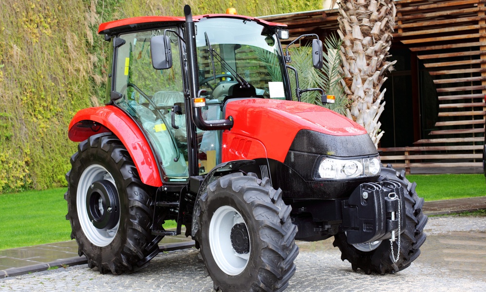 A red compact tractor sits on a gray concrete path. A large tree and a grassy area appear behind the tractor.