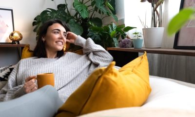 A woman sits on her couch in her home, looks out her window, and smiles. She is drinking something in a yellow mug.