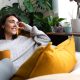 A woman sits on her couch in her home, looks out her window, and smiles. She is drinking something in a yellow mug.