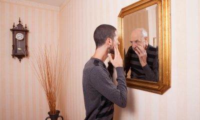 A young man looks at himself in the mirror with his hand on his face, but looking back is an older version of himself.