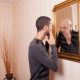 A young man looks at himself in the mirror with his hand on his face, but looking back is an older version of himself.