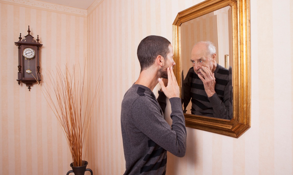 A young man looks at himself in the mirror with his hand on his face, but looking back is an older version of himself.