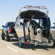 A black pickup truck towing a silver and black boat on a multi-lane highway surrounded by open fields.
