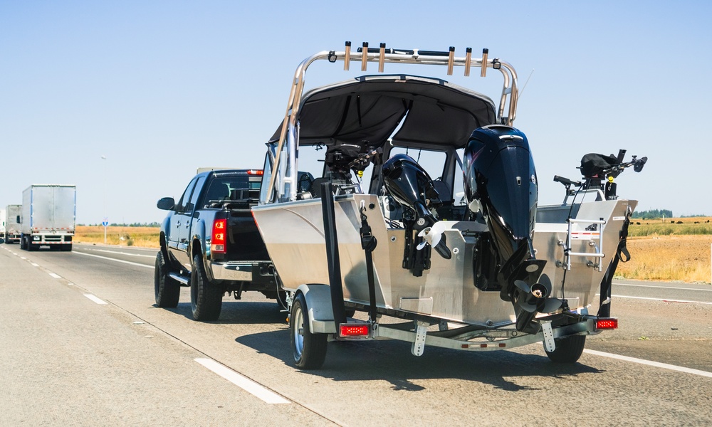 A black pickup truck towing a silver and black boat on a multi-lane highway surrounded by open fields.