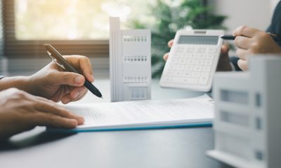 A person holding a pen prepares to sign a document while another person holds a calculator. Model buildings sit on the table.