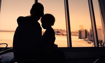 A shadow silhouette of a woman and a younger child sitting at an airport. Behind them is a view of the tarmac.