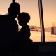A shadow silhouette of a woman and a younger child sitting at an airport. Behind them is a view of the tarmac.
