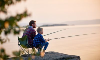 A father and son sitting on camping chairs and fishing on a boulder next to a calm lake at sunset.