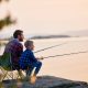 A father and son sitting on camping chairs and fishing on a boulder next to a calm lake at sunset.