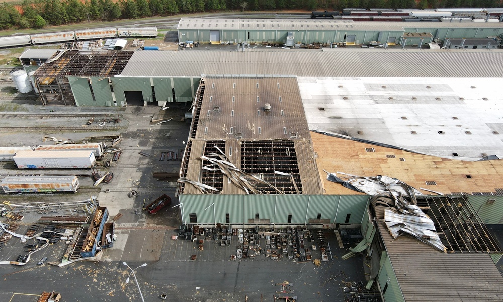 A aerial view of a large, green commercial building with the roof torn off in the aftermath of a tornado.