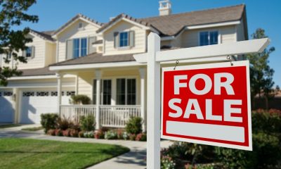 A red and white FOR SALE sign in front of a white house with a gray roof. The house is out of focus.