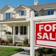 A red and white FOR SALE sign in front of a white house with a gray roof. The house is out of focus.