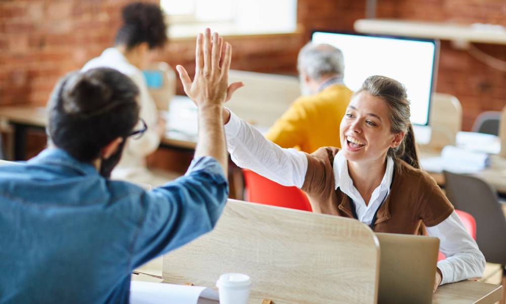 A young woman wearing a brown sweater smiles as she gives a high-five to a bearded man sitting across a desk.