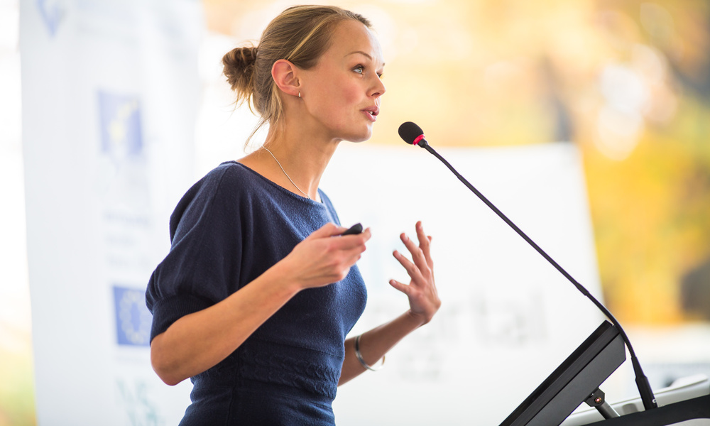 A blonde woman in a blue top and silver jewelry stands at a podium, speaking into a long microphone.