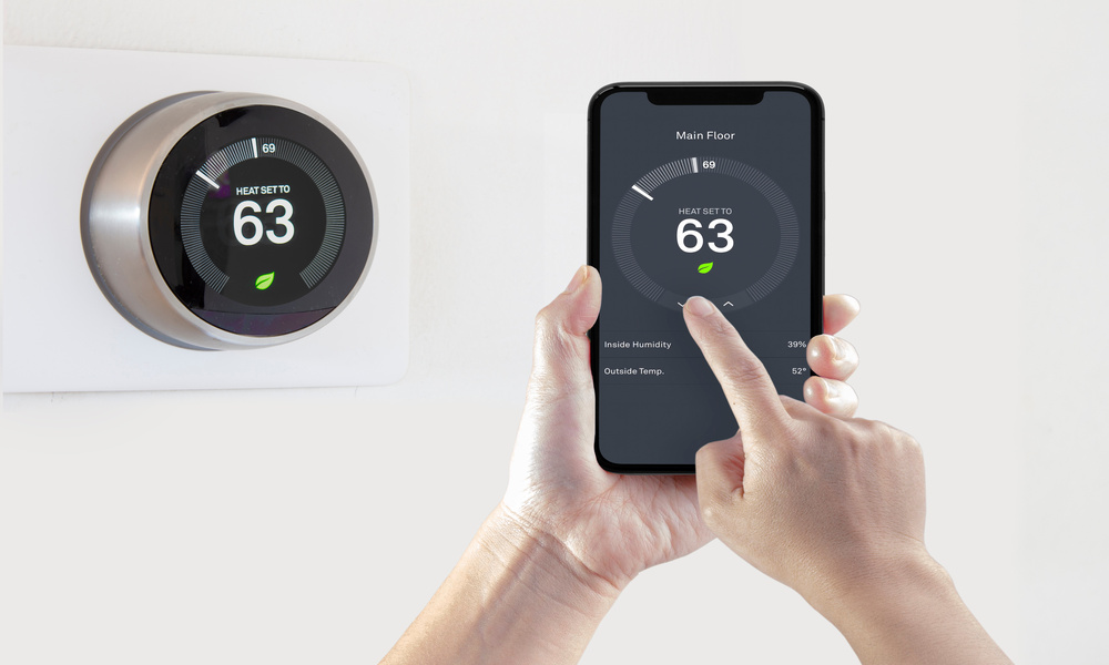 A round, digital thermostat displays a dial and the number "63." A person holds a smartphone that displays the same image.