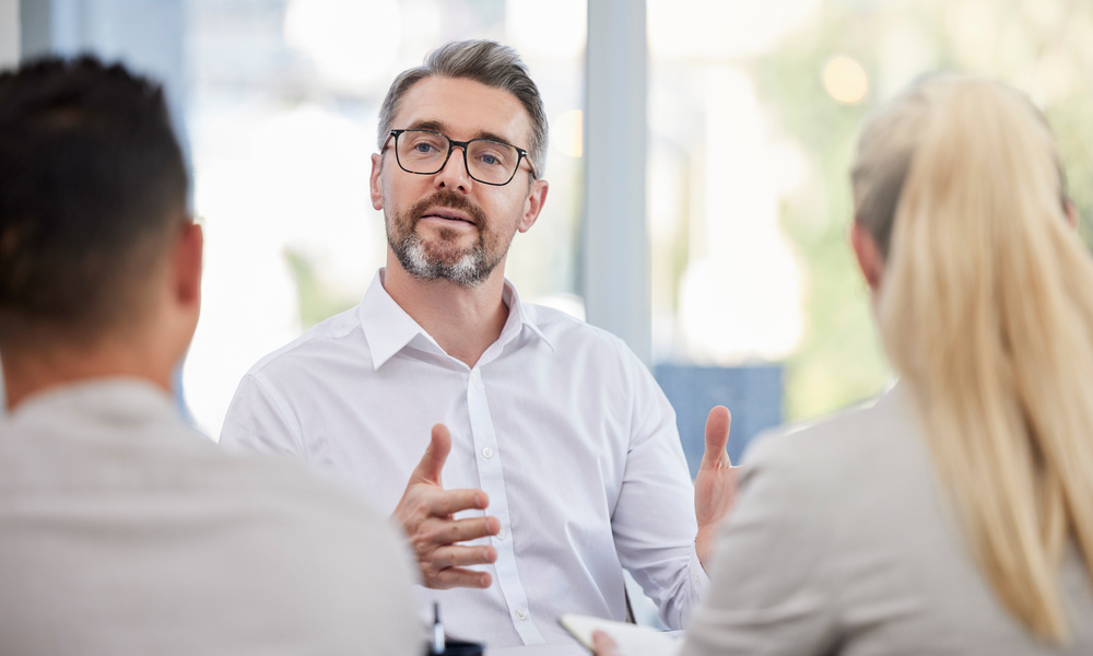 A businessman wearing glasses talks to two people in front of him. The three sit in his office, which has a window.